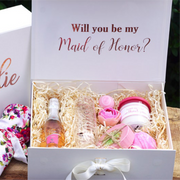A photo of a light ivory-colored "Will you be my bridesmaid?" box with a magnetic and ribbon closure. The inside flap has a message written in rose gold script font. The box contains various items, including a small bottle of champagne, a stemless champagne flute, a scented candle and soap, and a handwritten note. The items are arranged neatly inside the box with natural box filler and a floral robe. The overall aesthetic is elegant and feminine.